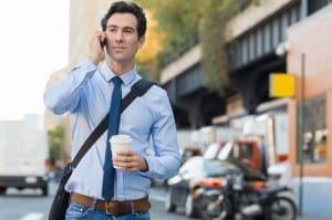 Happy businessman talking on the mobile phone. Portrait of a young businessman communicating over cell phone while holding a paper cup and walking on the road. A young trendy smiling guy thinking at smartphone and looking away while going to work.